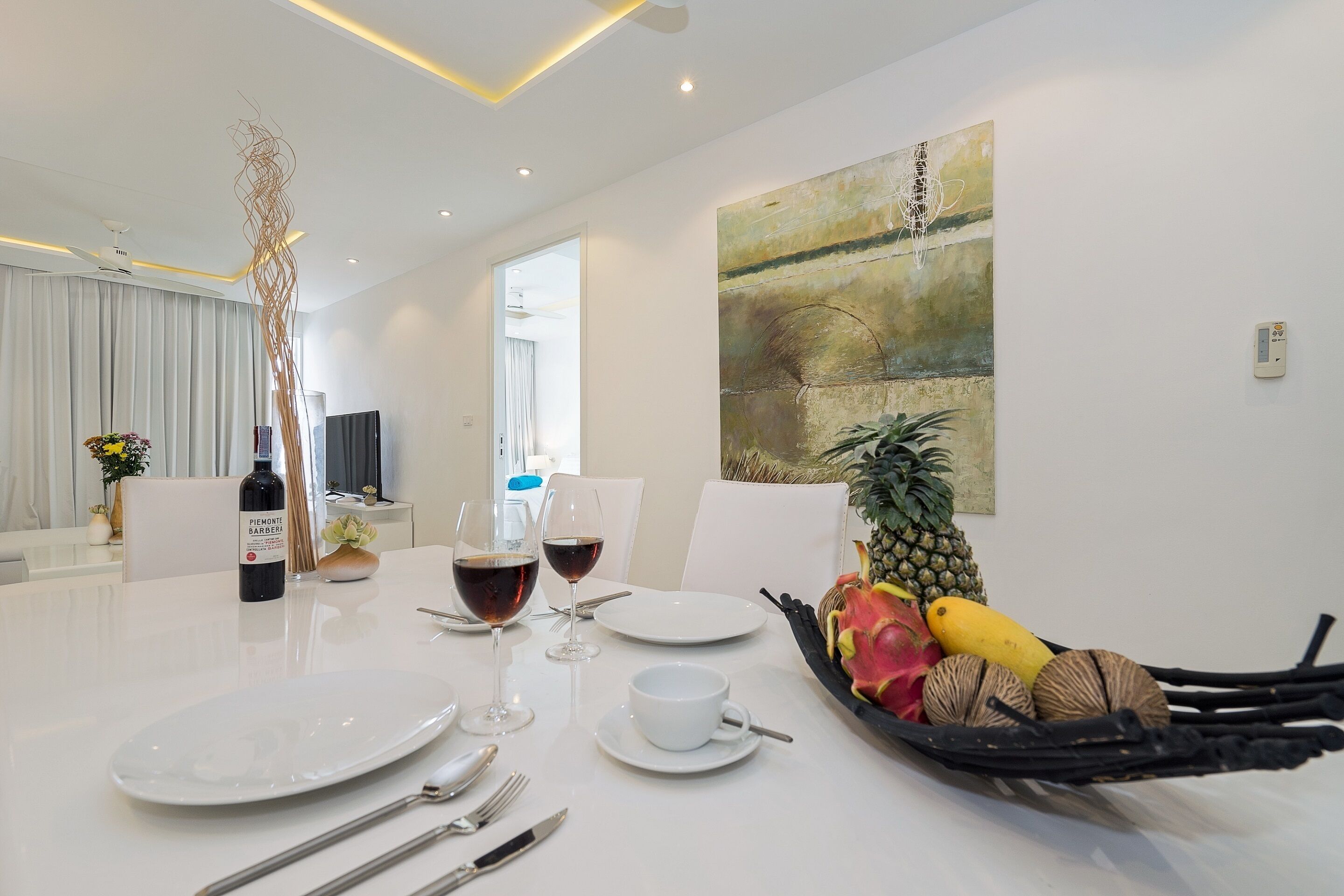 A modern dining area with a white table set for two, featuring wine glasses, plates, and a fruit basket.