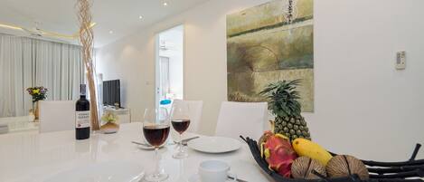 A modern dining area with a white table set for two, featuring wine glasses, plates, and a fruit basket.