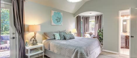 A bedroom with a large bed, a nightstand, a lamp, and a view of the outdoors.