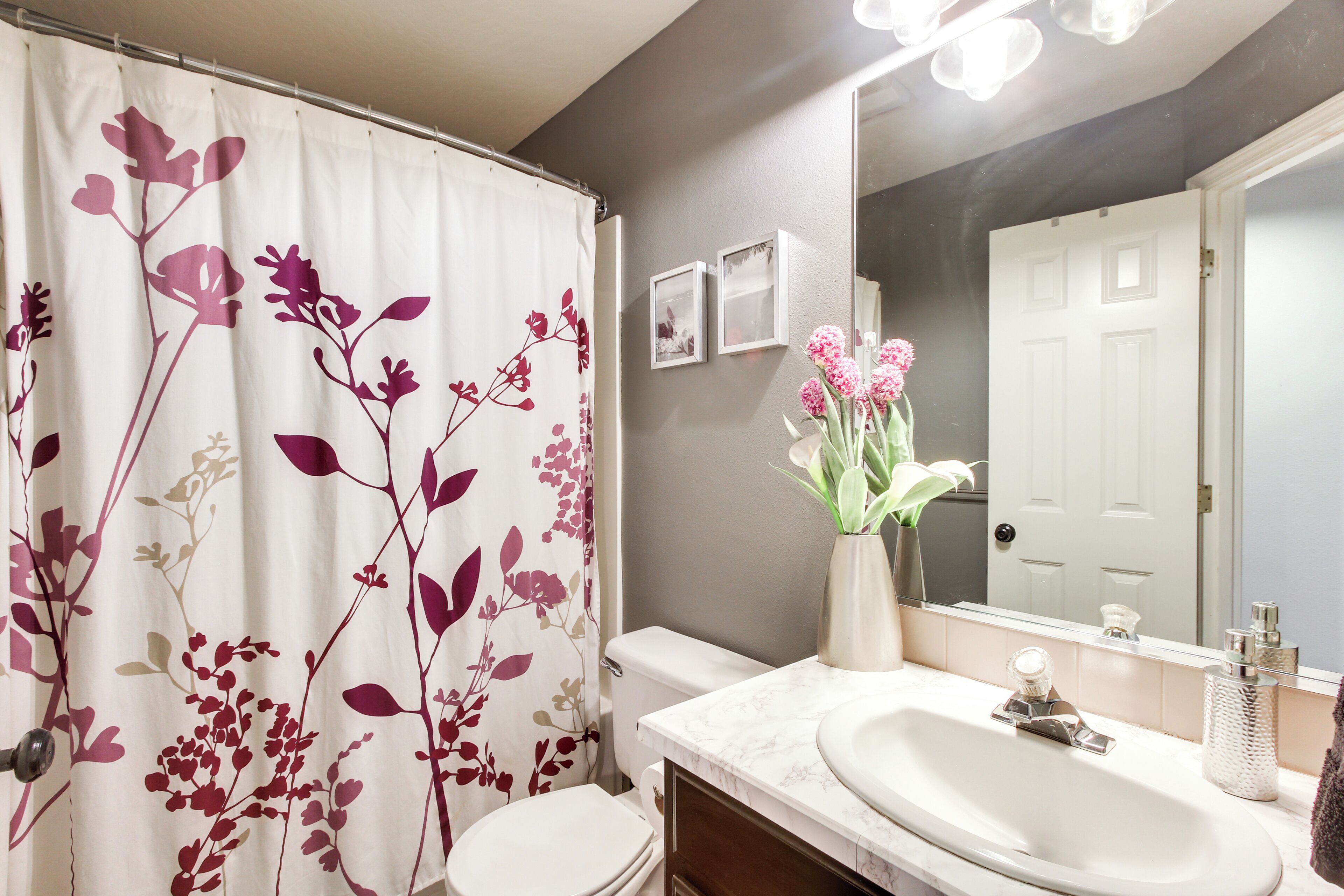 Ein Badezimmer mit Toilette, Waschbecken mit Marmorplatte, einer Duschvorhang mit Blumenmuster, einer Vase mit rosa Blumen und einem Spiegel über dem Waschbecken.