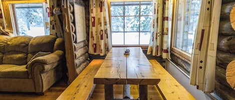 A log cabin interior with a wooden table and benches, a sofa, and large windows with curtains.