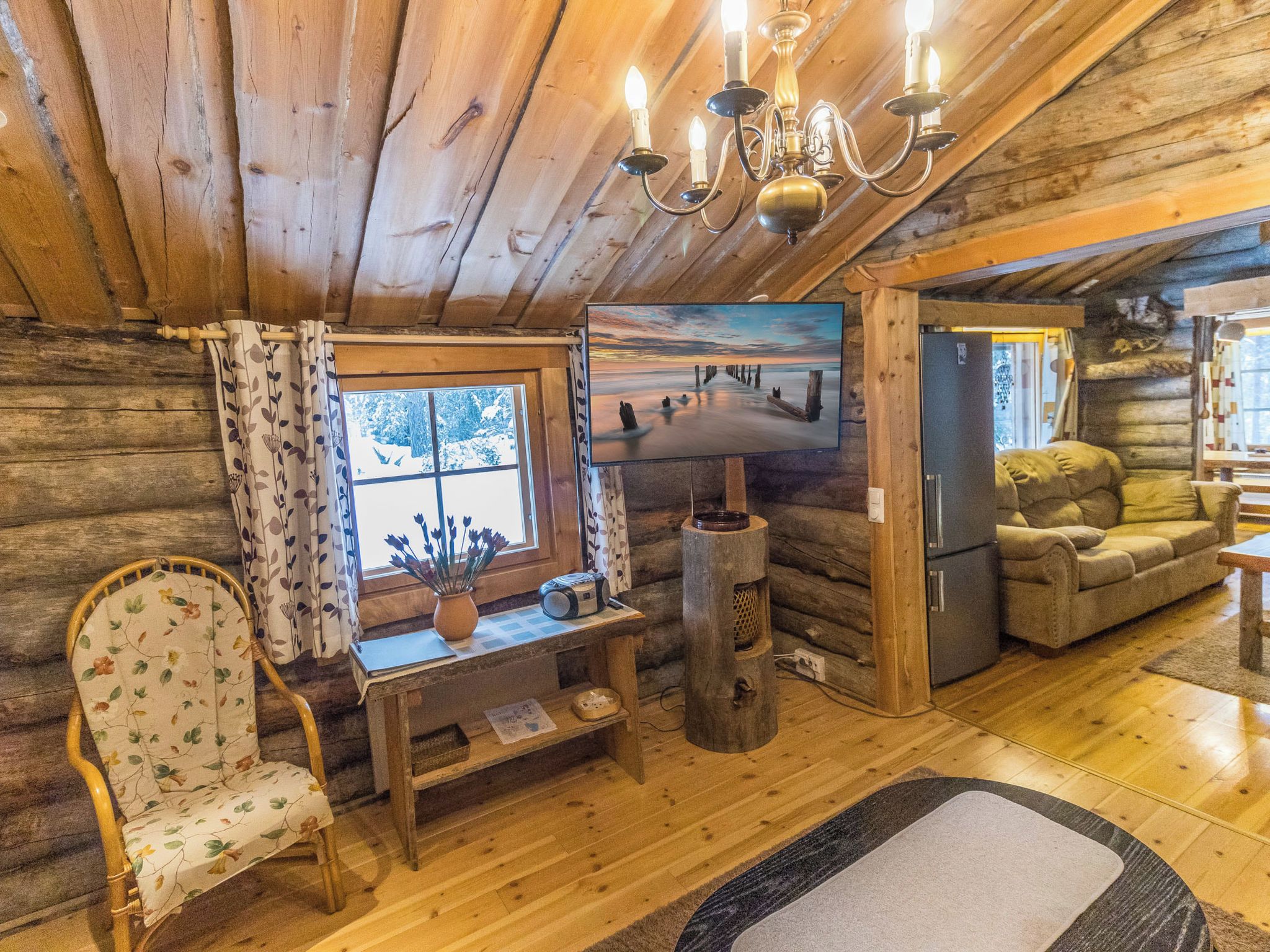 A cozy log cabin interior with a wooden ceiling, a television mounted on the wall, a sofa, a chair, and a small table with a vase and a radio.