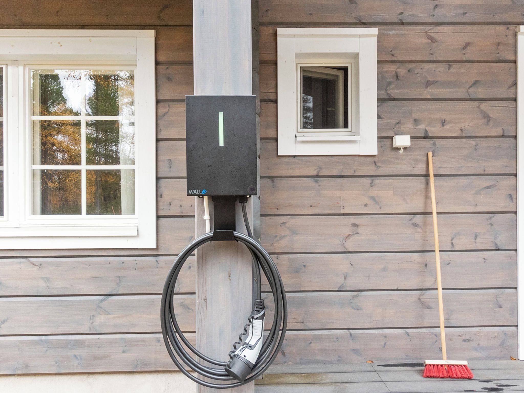 A utility box mounted on a wooden wall with a coiled cable and a broom leaning against the wall.