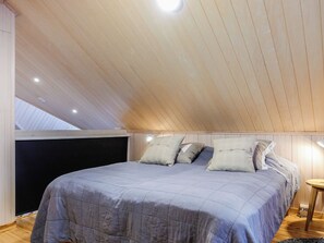 Une chambre avec un plafond en pente, un lit recouvert d’une couette grise et deux lampes de chevet.