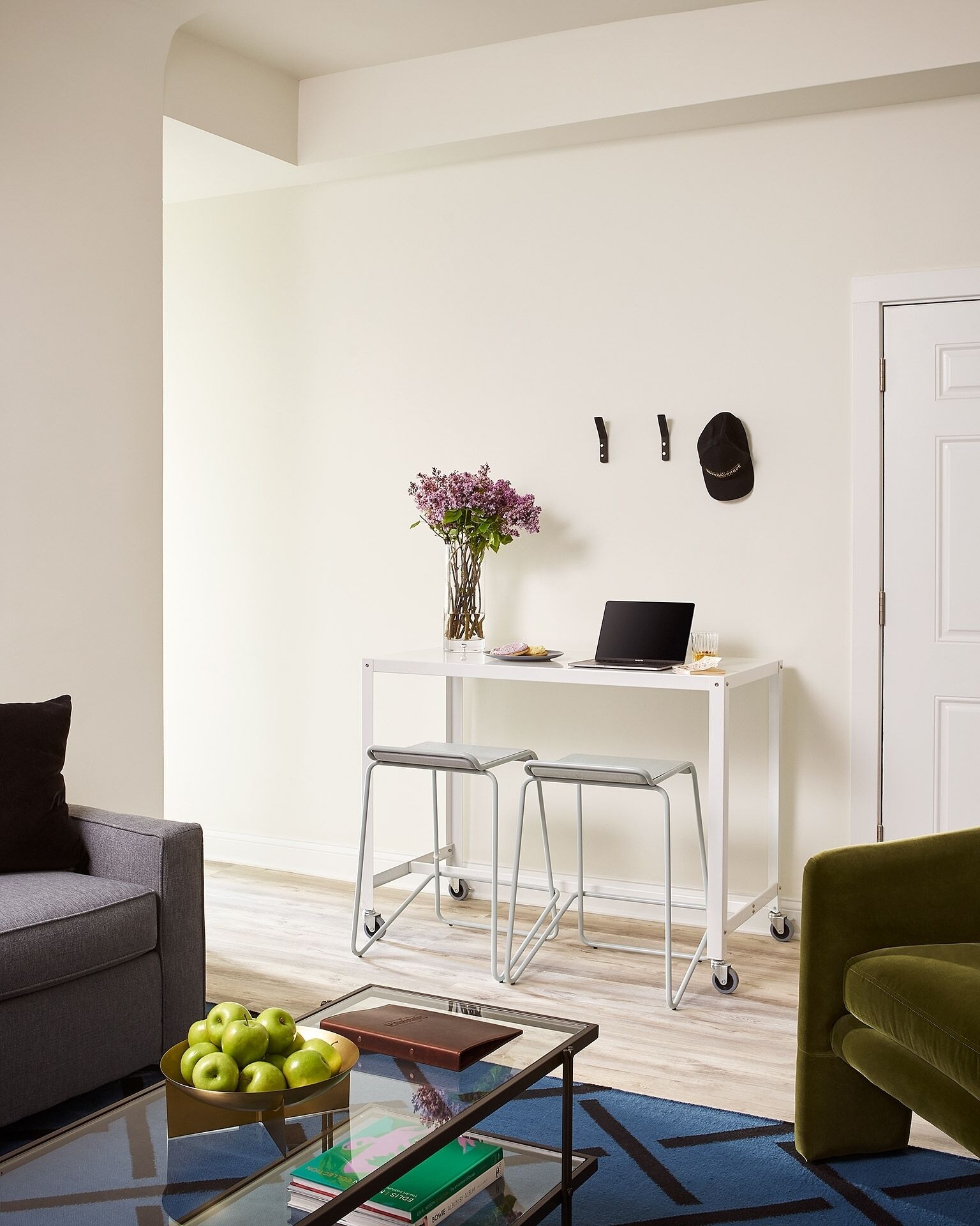 A modern living room with a grey sofa, a green armchair, a glass coffee table with books and apples, a white desk with a laptop and a vase of flowers, and a white door.