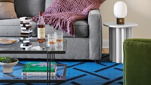 Un salon avec un canapé, une table basse sur laquelle se trouvent des livres et une bouteille, une petite table ronde avec une lampe, et un tableau encadré accroché au mur.