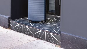 Une valise bleue sur un sol carrelé, devant un bâtiment avec une porte noire et un appareil numérique fixé au mur.