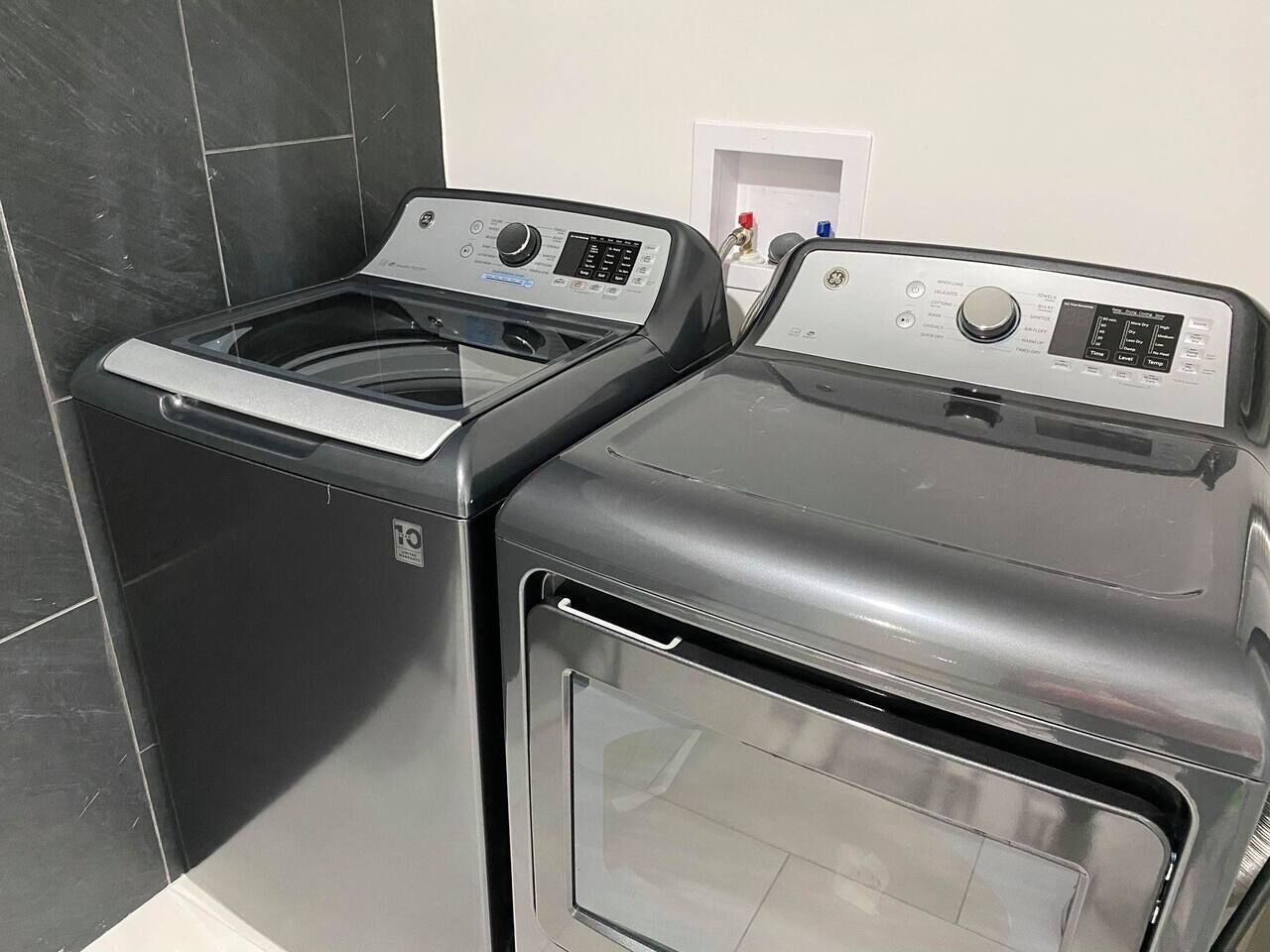 Une petite buanderie avec deux appareils superposés, un lave-linge à chargement frontal et un sèche-linge à chargement par le haut, le tout adossé à un mur carrelé.