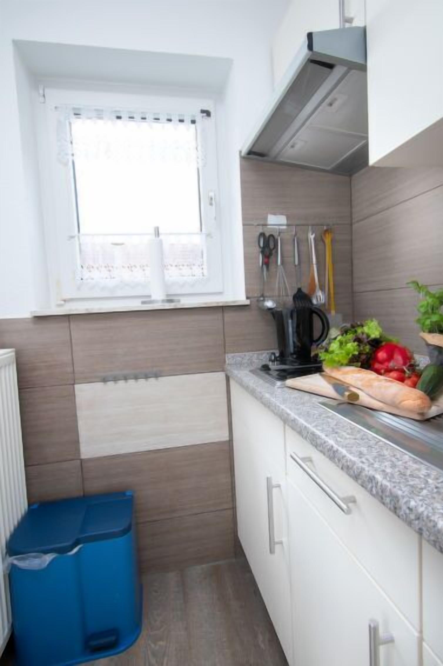 A small kitchen with a window, a blue trash bin, and a countertop with vegetables and a knife.