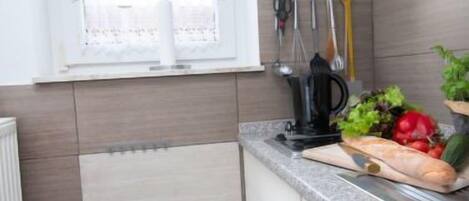 A small kitchen with a window, a blue trash bin, and a countertop with vegetables and a knife.