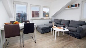 A modern, well-lit living room with a grey sofa, a dining table with chairs, and a small white coffee table. There are large windows providing natural light and a view of the outside. - Leuchtturm by Interhome (Norddeich)