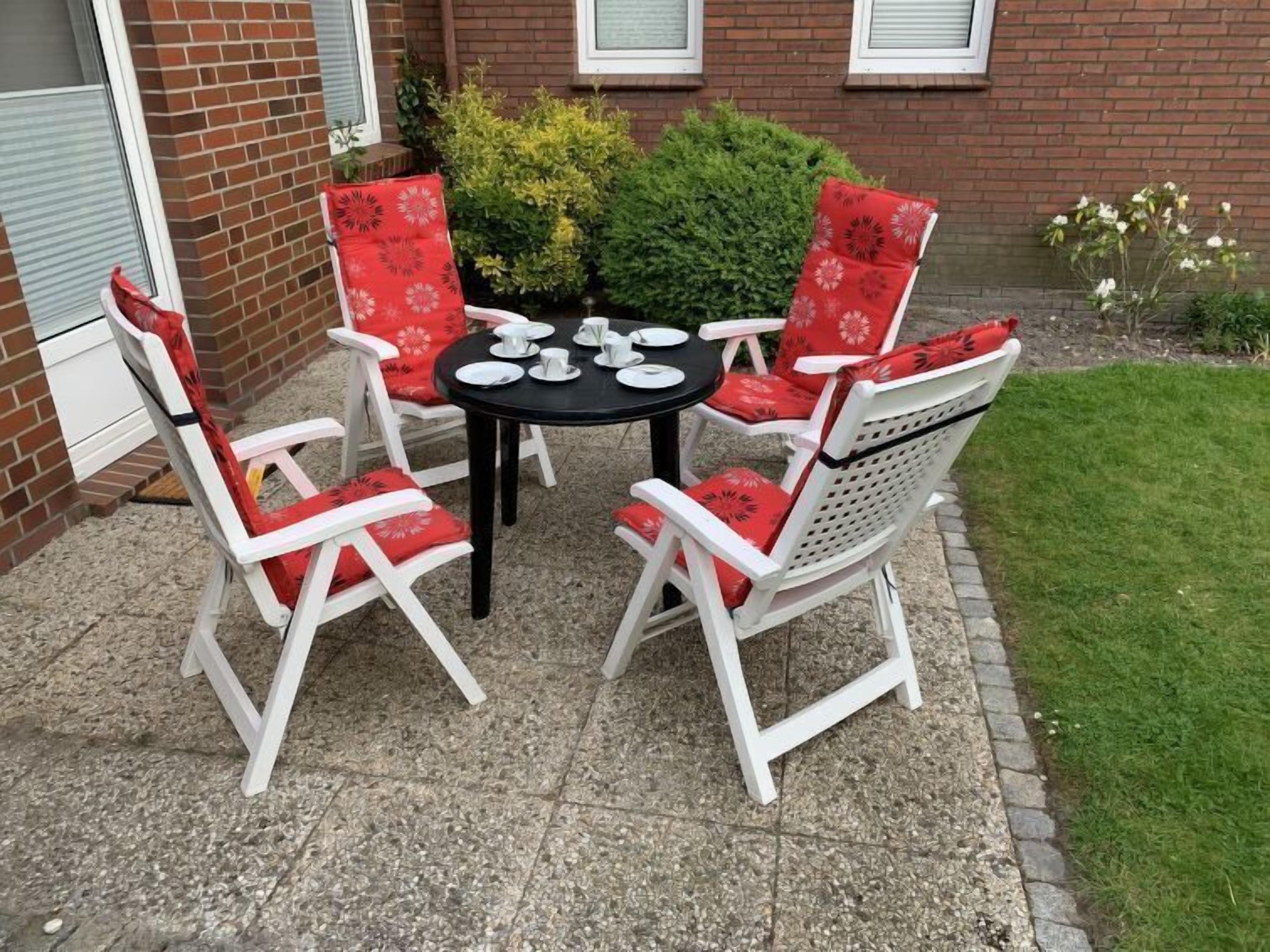 A patio with a round table and four white chairs with red cushions, set against a brick wall and a grassy area.