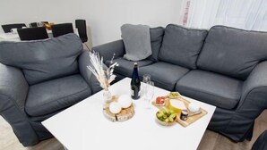 Un salon moderne avec un canapé en cuir gris, une table basse blanche et un petit coin repas comprenant une planche en bois et divers aliments.
