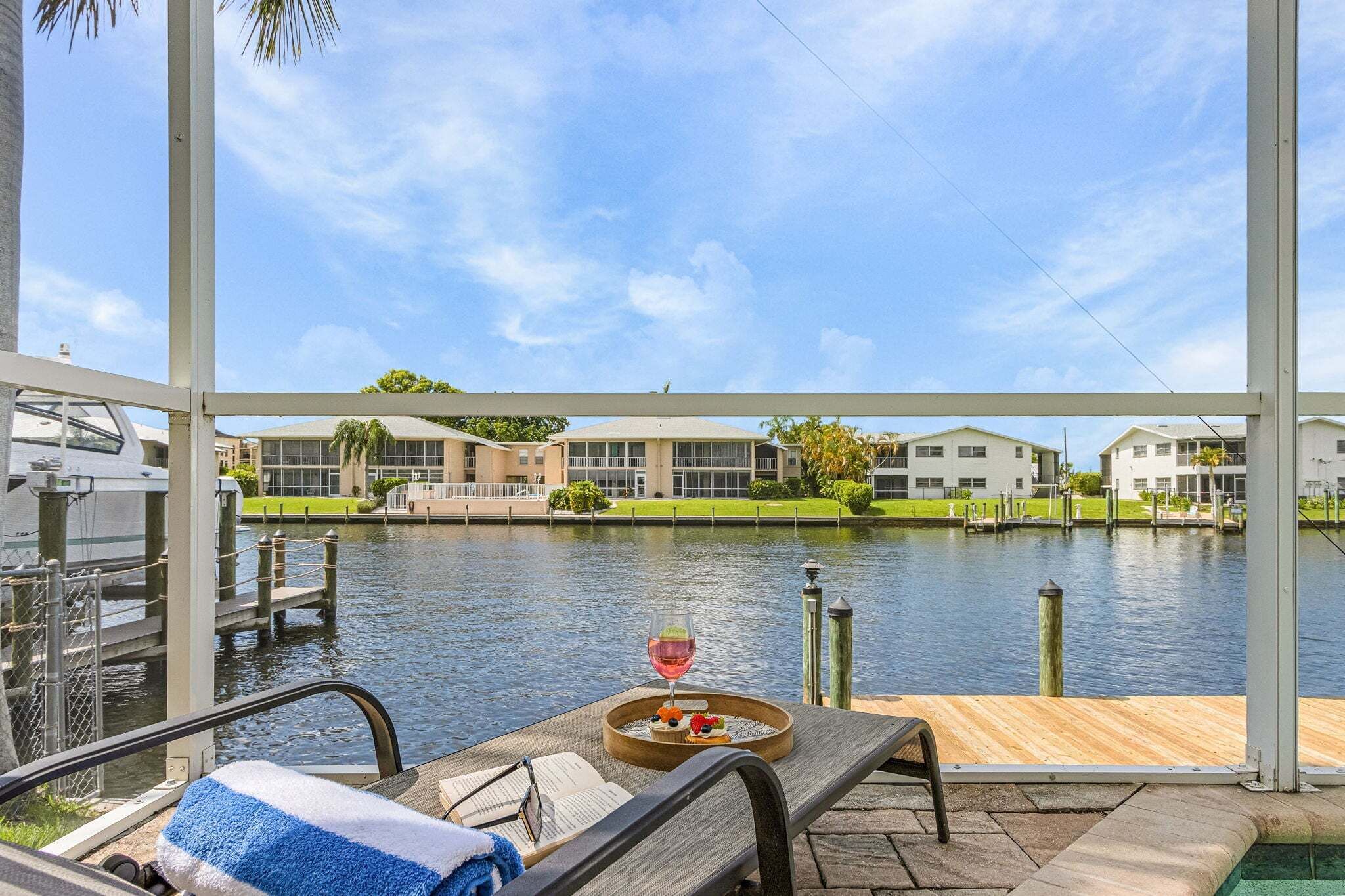 A waterfront view with a covered seating area, a dining table with a drink and snacks, and a pool in the background.