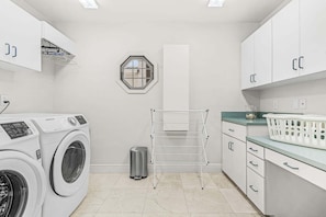 A laundry room with a washing machine, dryer, drying rack, trash bin, and storage cabinets.