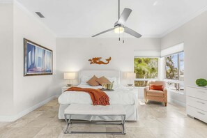 Un dormitorio con cama, mesitas de noche, ventilador de techo, ventana con vistas y un cuadro en la pared.
