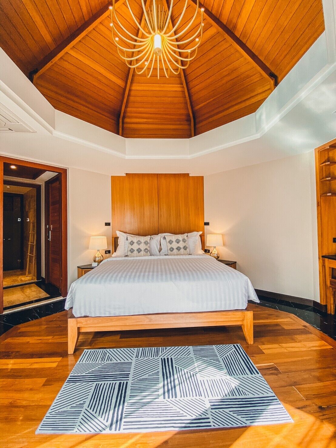 Un dormitorio con techo de madera, cama con cubrecama blanco, dos lámparas de noche y una alfombra estampada.