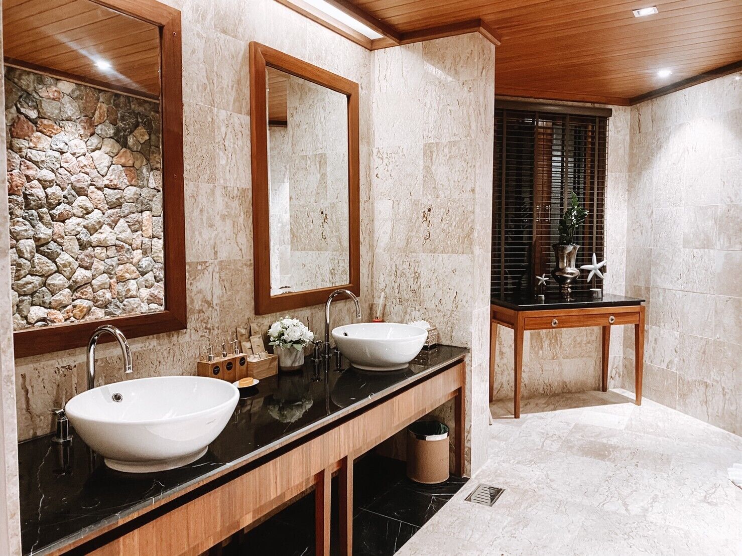 Une salle de bain avec deux lavabos, un meuble-lavabo en bois, un mur en pierre et une fenêtre avec des stores.