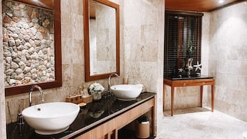 A bathroom with two sinks, a wooden vanity, a stone wall, and a window with blinds.