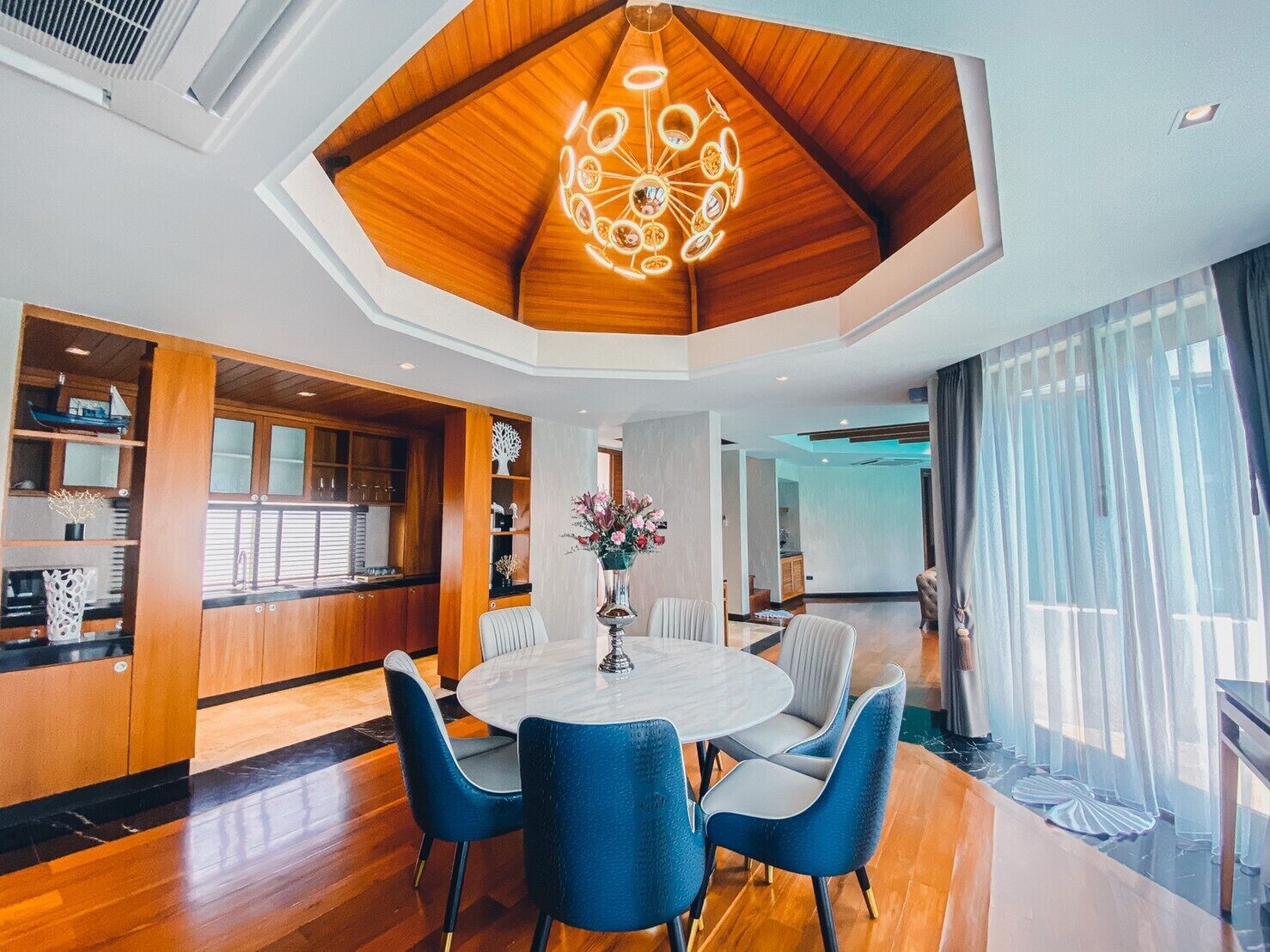 A modern dining area with a round table and blue chairs, a wooden ceiling, and a view of a kitchen and living space.