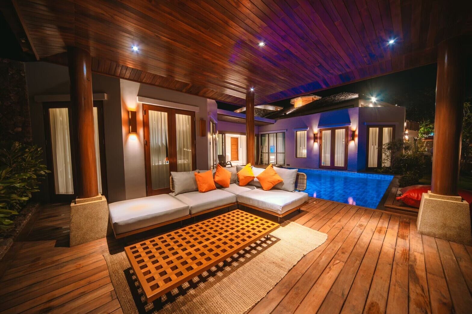 A modern outdoor living area with a wooden deck, a sectional sofa with orange cushions, and a pool.