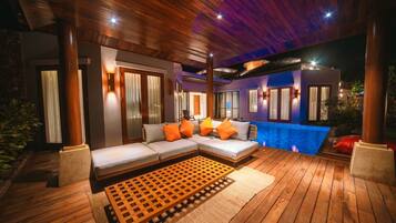 A modern outdoor living area with a wooden deck, a sectional sofa with orange cushions, and a pool.