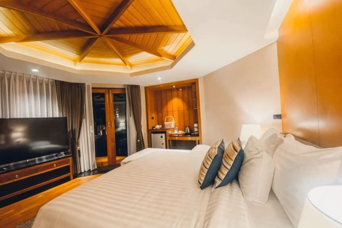 A hotel room with a large bed, a TV, a wooden ceiling, and a small kitchenette.