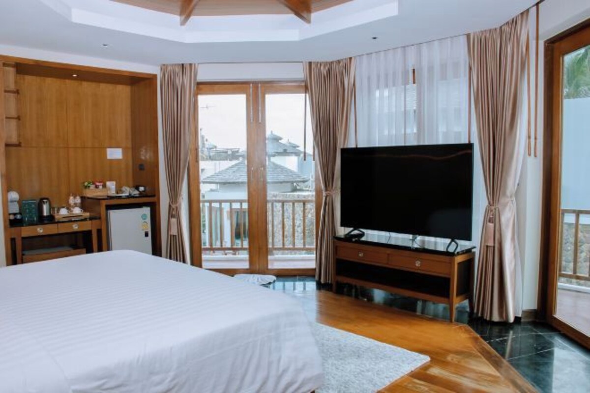 A hotel room with a large bed, a TV on a wooden stand, a desk with a computer, and a balcony with curtains.