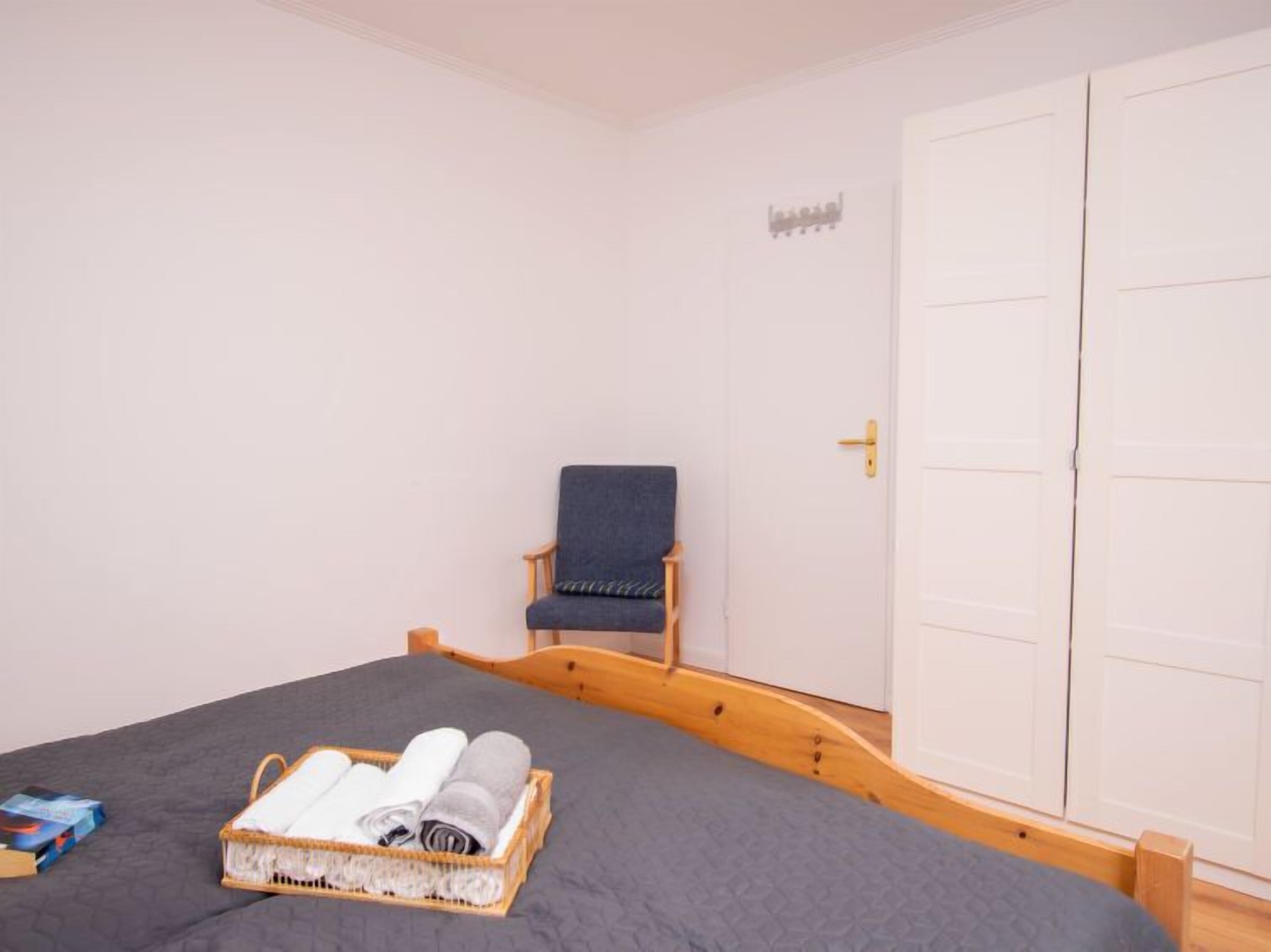 A bedroom with a bed, a chair, a wardrobe, and a basket of towels.