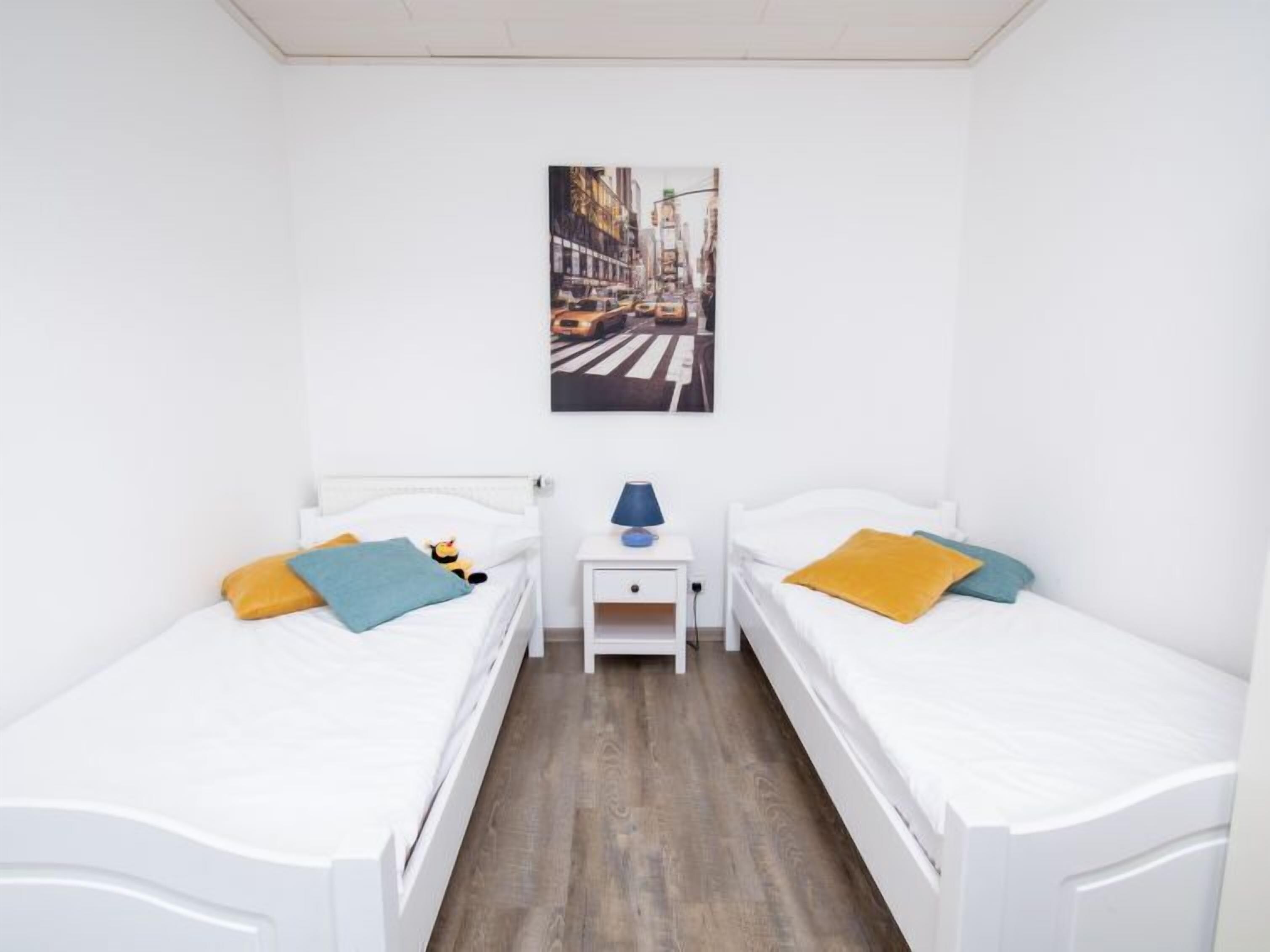 A room with two single beds, a nightstand with a lamp, and a painting of a city street on the wall.