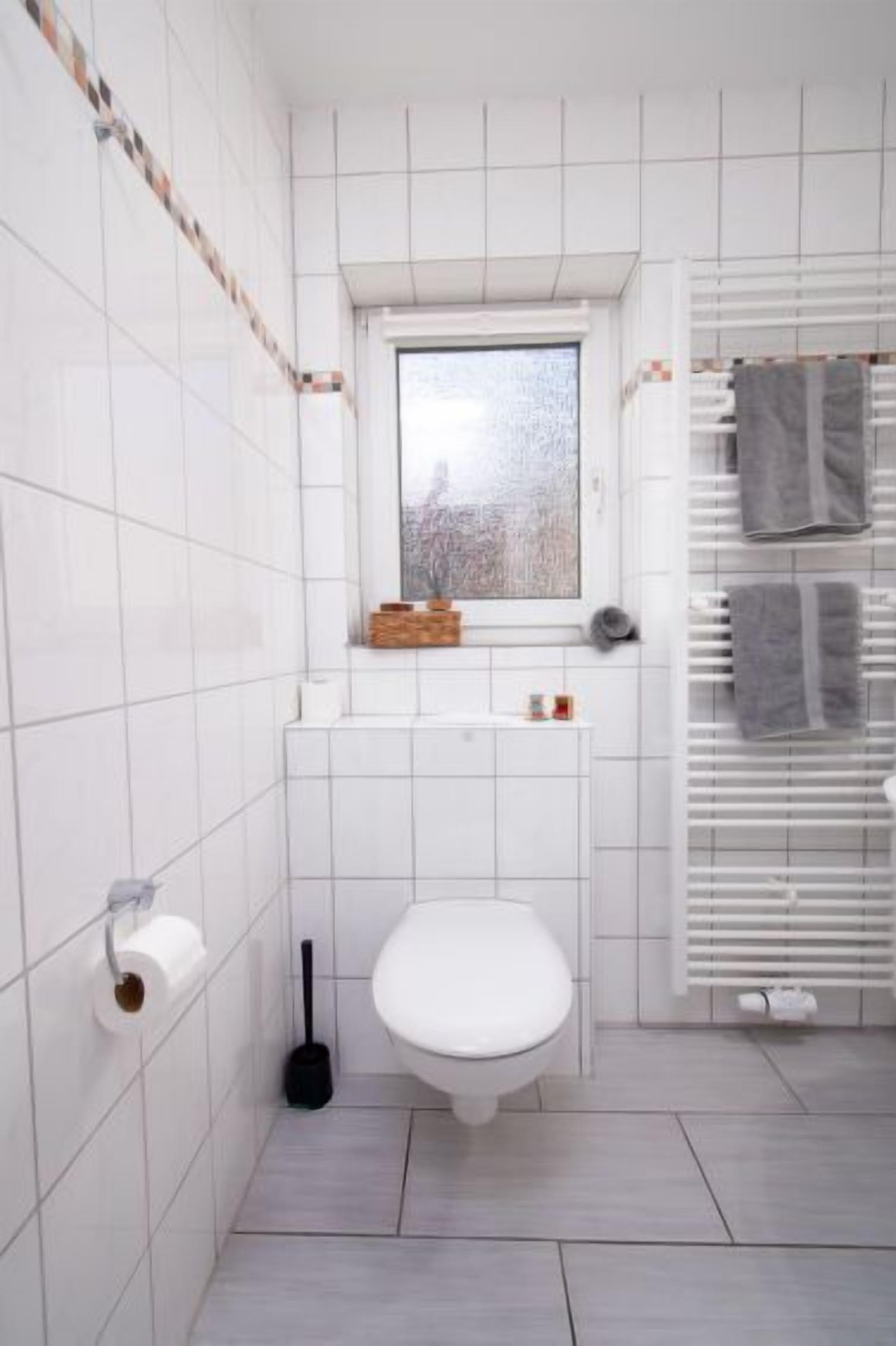 Une salle de bain avec des carreaux blancs, des toilettes, une fenêtre, un porte-serviettes et un radiateur.
