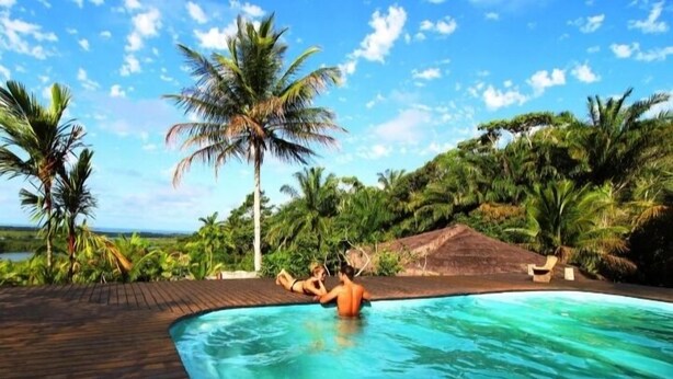 Una piscina con dos personas en el agua, rodeada de palmeras y una terraza de madera.