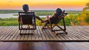 Dos personas sentadas en sillas de madera mirando hacia un río, con un atardecer al fondo.