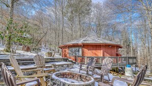 Un terrazzo in legno con poltrone Adirondack e un braciere in pietra circondato dalla neve, con una baita di legno sullo sfondo.