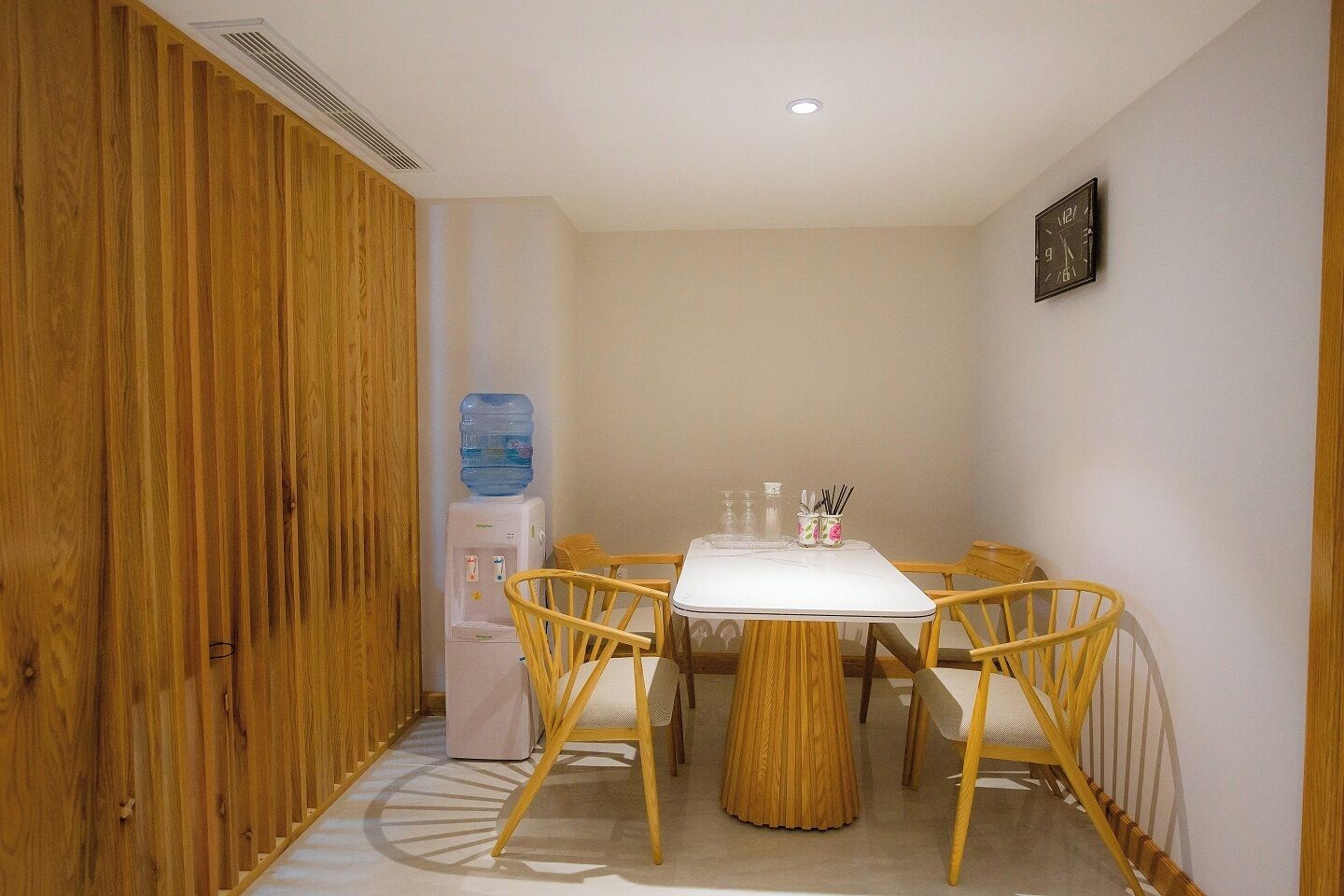 A small dining area with a white table, four wooden chairs, a water dispenser, and a clock on the wall.