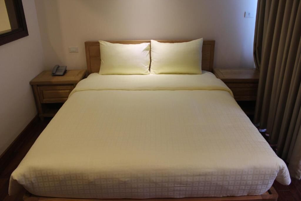 Un llit ben fet amb un estovat estampat i dos almoinos, una tauleta de nit amb un telèfon, i una cortina mig tancada.