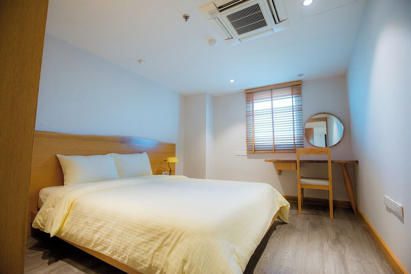 A hotel room with a bed, a desk, a mirror, and a window with blinds.