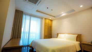 A hotel room with a bed, a bedside table, a nightstand, a window with curtains, and a ceiling with an air conditioning unit.