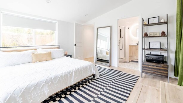 Ein modernes Schlafzimmer mit einem großen Bett, einem schwarz-weiß gestreiften Teppich und Blick auf ein Badezimmer und eine Küche durch eine offene Tür.