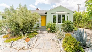 Ein eingeschossiges Haus mit einer gelben Tür, umgeben von einem Garten mit verschiedenen Pflanzen und einem Kiesweg.