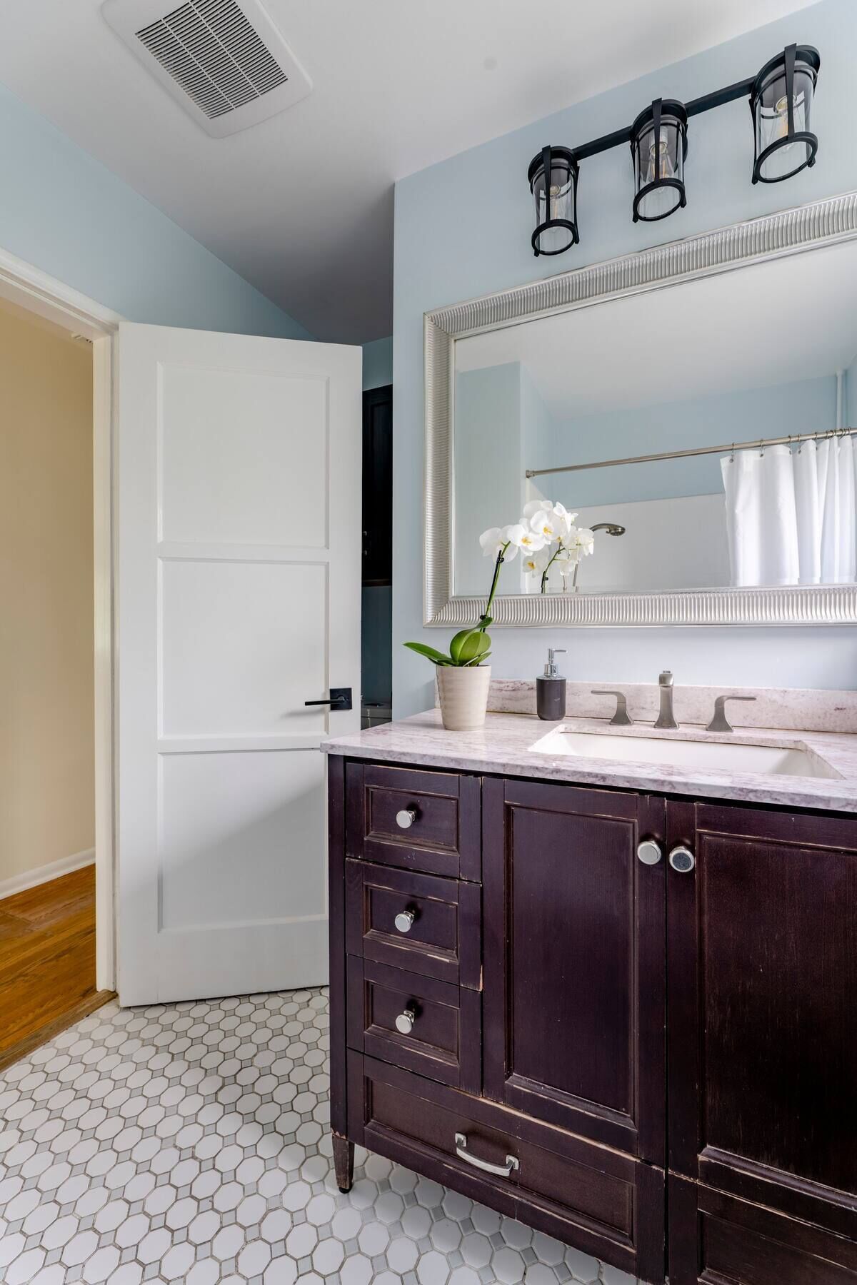A bathroom with a white door, a dark wooden vanity with drawers, a large mirror, and a hexagonal tiled floor.