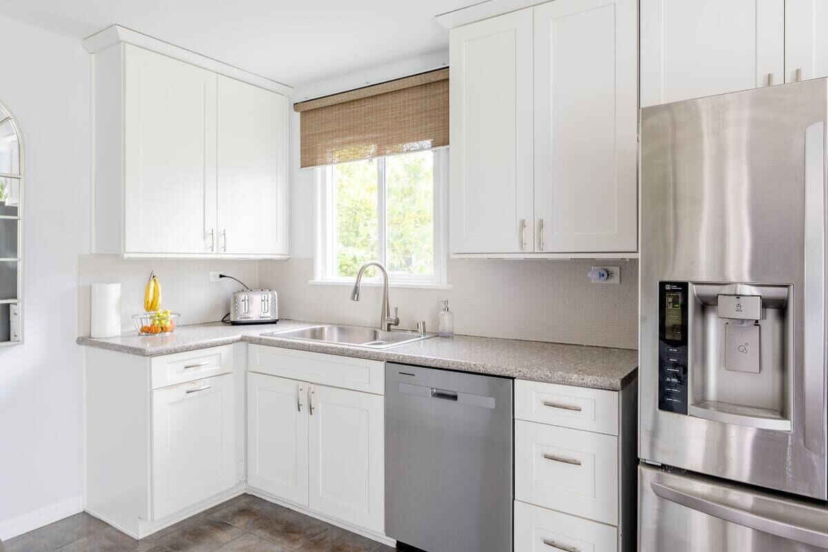 A modern kitchen with white cabinets, a stainless steel refrigerator, a dishwasher, and a window with a beige blind.