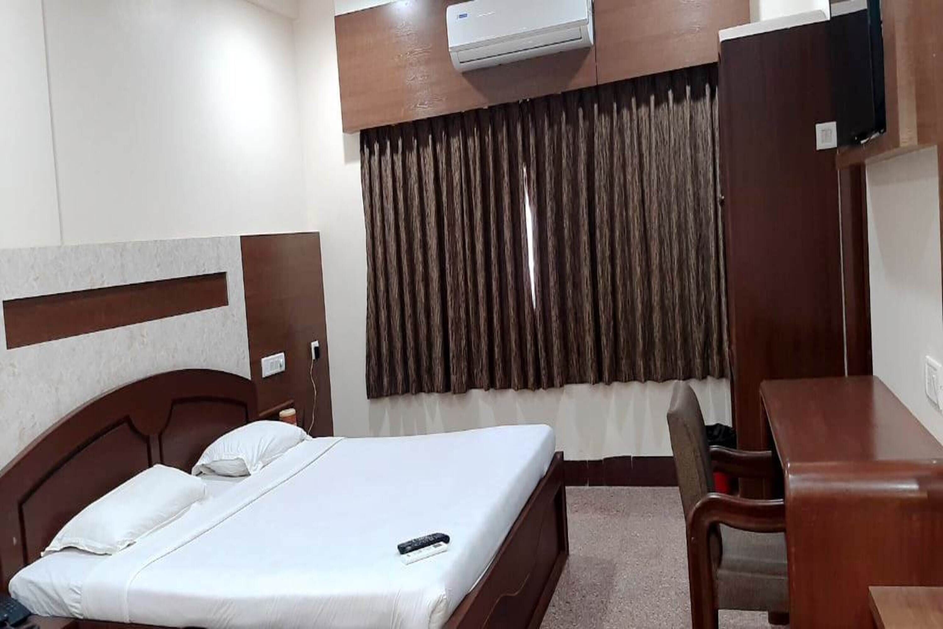 A hotel room with a bed, a desk, a chair, an air conditioning unit, and a window with curtains.