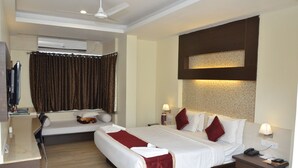 A hotel room with a bed, a desk, a chair, a TV, and an air conditioning unit. - Hotel Adarsha Palace (Belagavi)