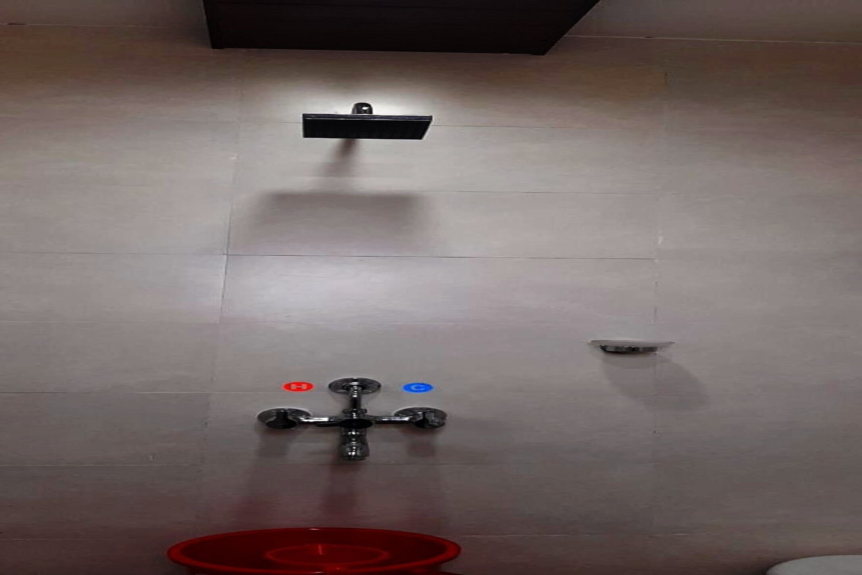 A bathroom with a wall-mounted faucet, a red trash bin, and a small sensor on the wall.