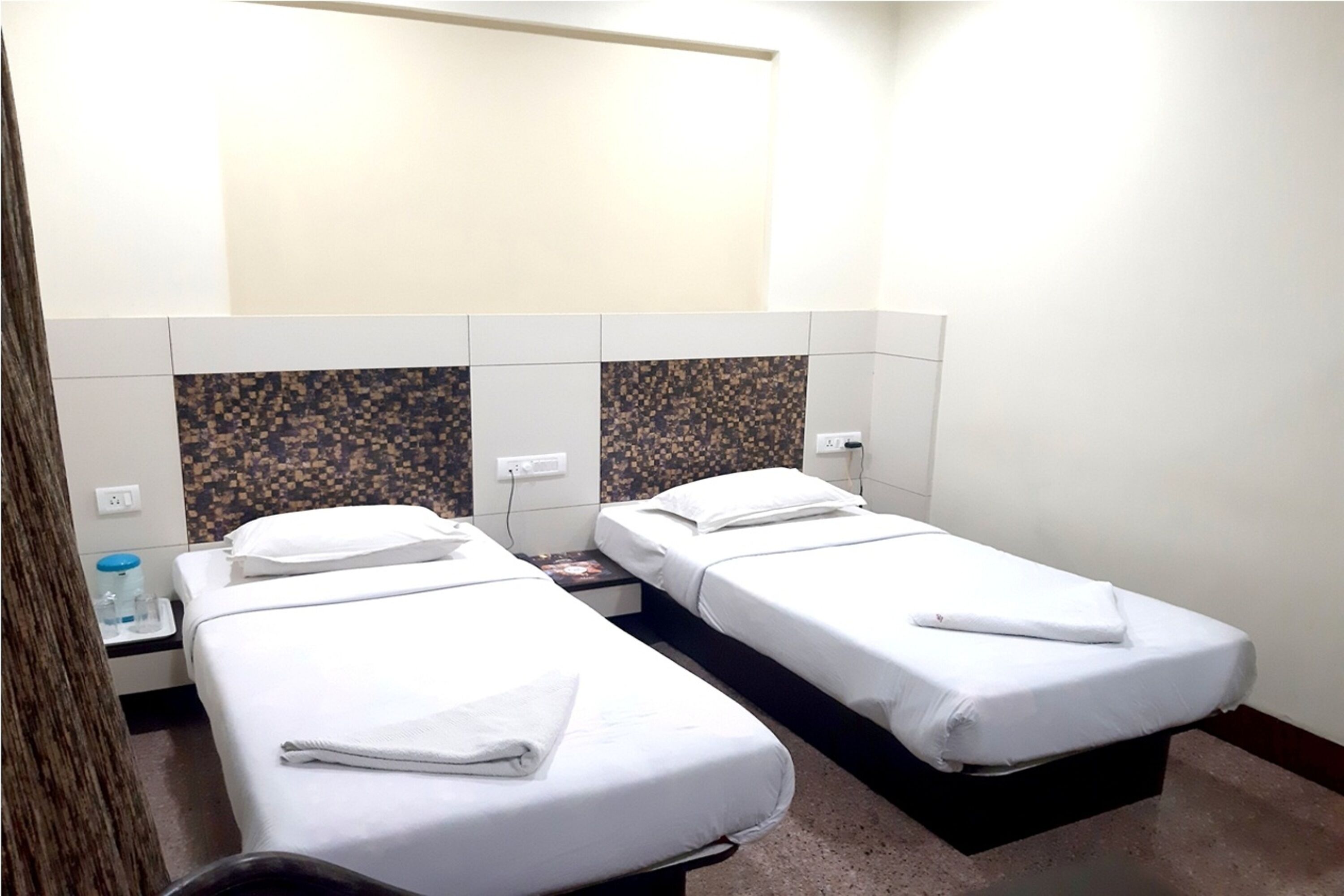 Een hotelkamer met twee enkele bedden, elk met een witte matras en kussen, een klein nachtkastje en een muur met een decoratief tegelpatroon.