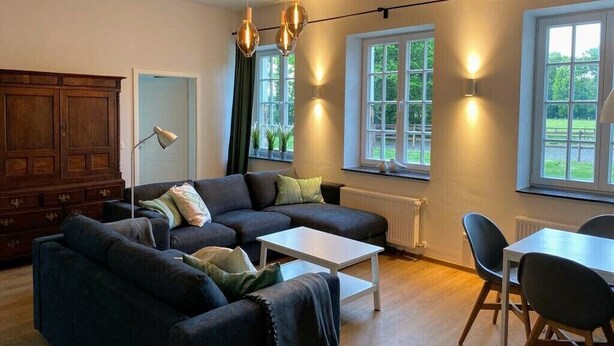 A modern living room with a sectional sofa, a white coffee table, and a dining table with chairs. There are large windows with views of greenery outside.