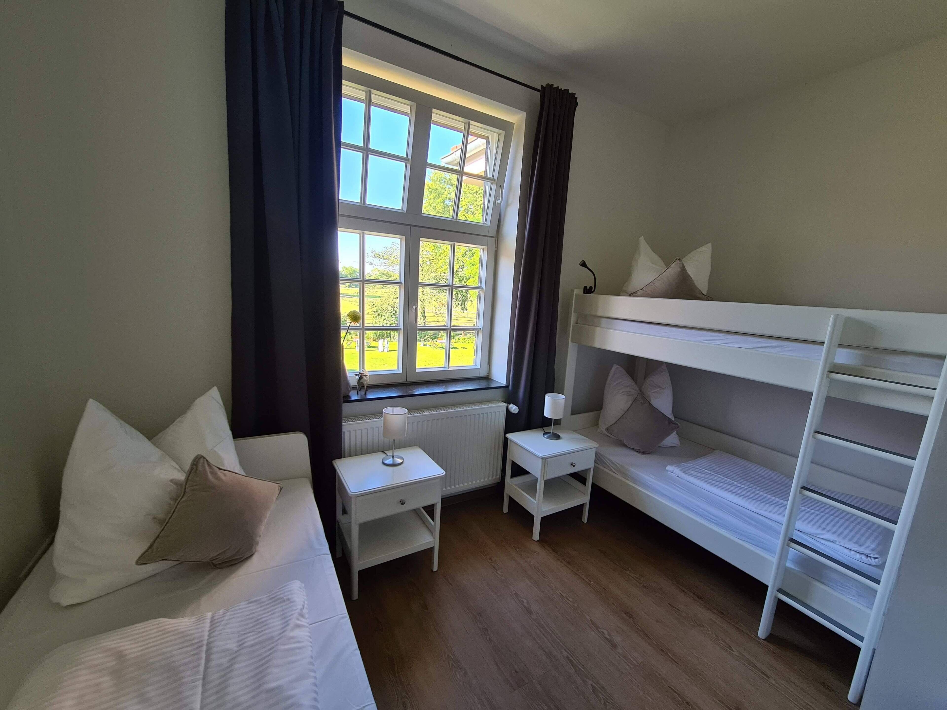 A bedroom with a bunk bed, a single bed, a bedside table with a lamp, and a window with curtains.
