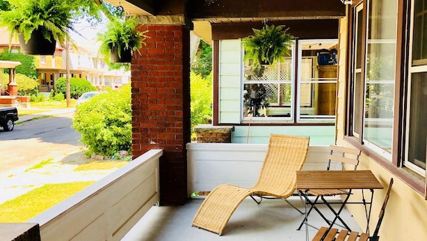Une terrasse extérieure couverte, avec une table et deux chaises, un mur de briques et une vue sur une rue bordée de maisons et de verdure.