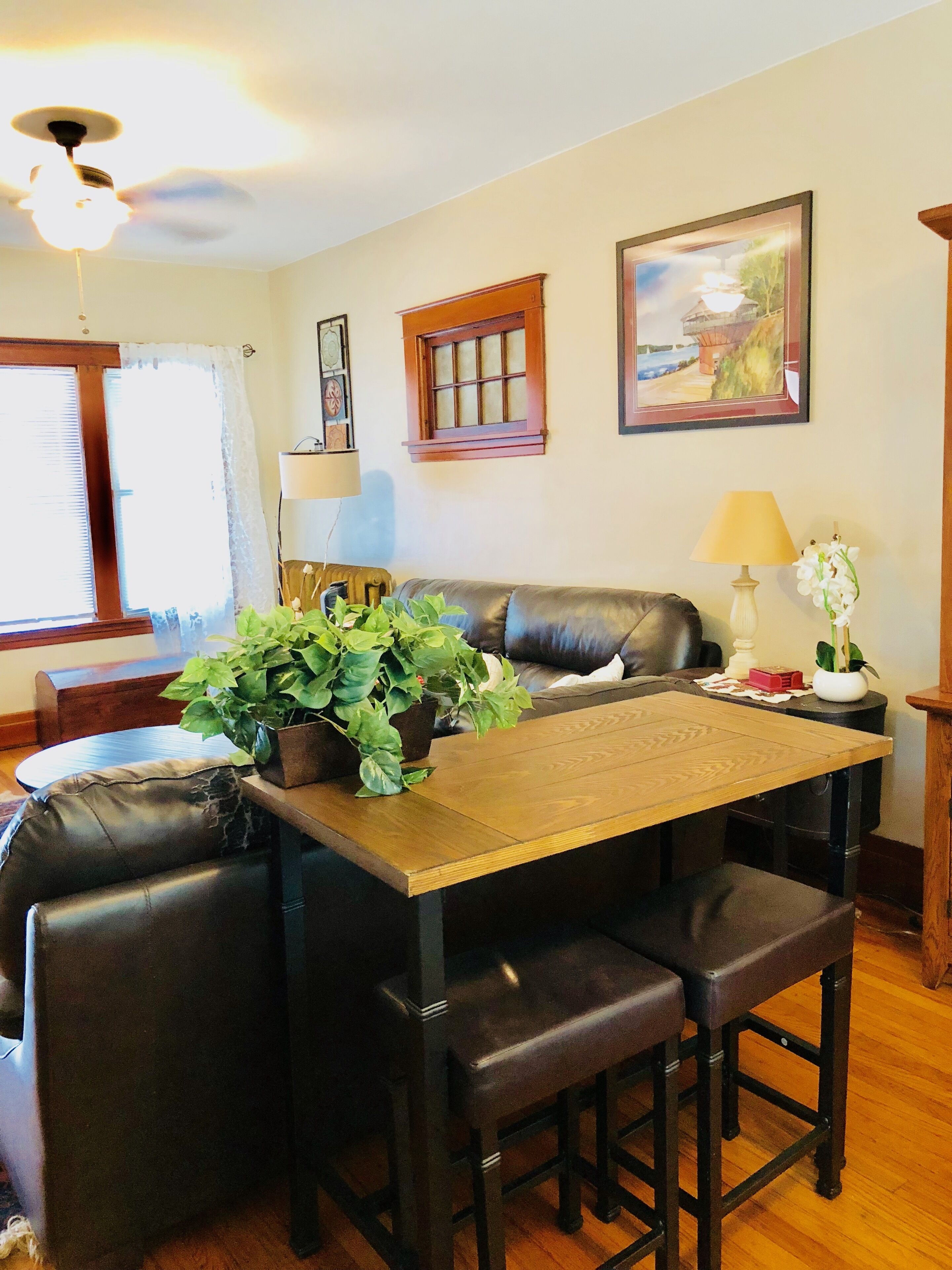 A dining area with a wooden table, black leather chairs, a potted plant, a ceiling fan, a window with blinds, and a framed picture on the wall.